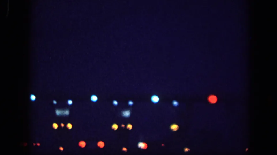 8mm home-movie frame circa 1963, possibly Memphis, Tennessee: scattered blue, amber, and red bokeh lights at night, with an amber cluster suggesting a rectangular illuminated sign or structure.