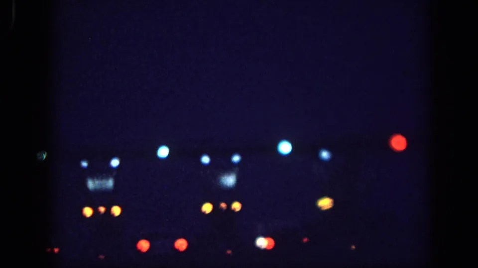 8mm home-movie frame circa 1963, possibly Memphis: blue-white, amber, and red out-of-focus light clusters at night, with a faintly visible rectangular illuminated structure in the background.