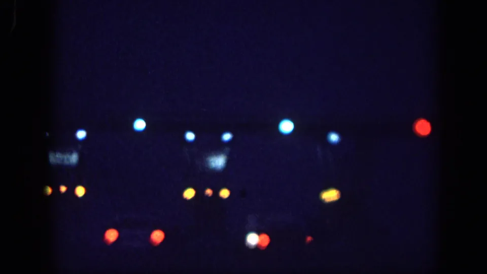 8mm home-movie frame circa 1963, possibly Memphis: a spread of blue, amber, and red bokeh lights consistent with commercial strip or venue lighting, with a faint illuminated structure background.