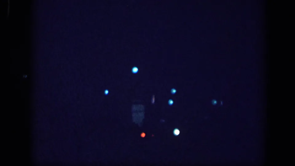 8mm home movie frame from a 1963 Memphis night drive; scattered cyan bokeh streetlights and a faint vehicle silhouette against deep blue darkness.