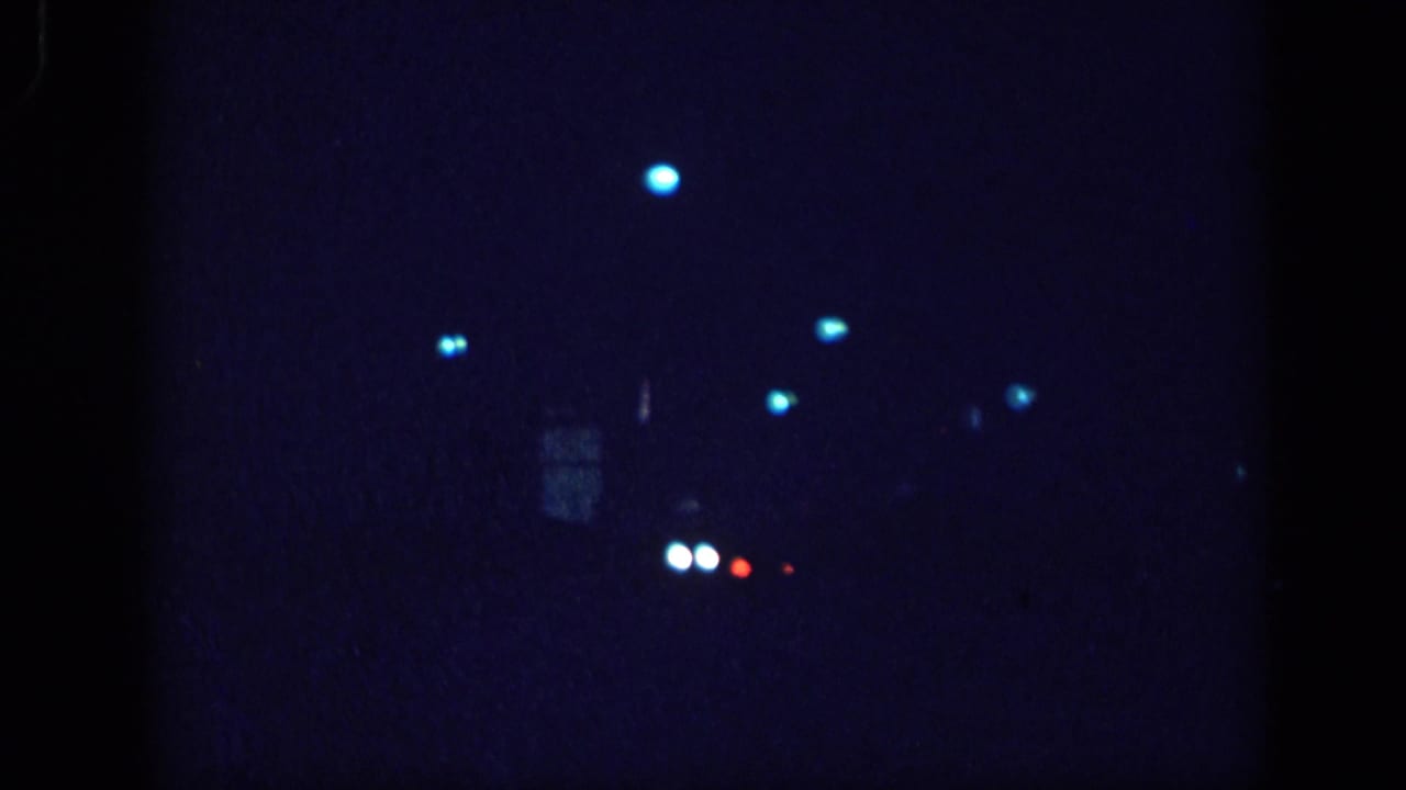Dark 8mm film frame with blue bokeh light points and a faint vehicle shape; paired white headlight bokeh dots visible at center.