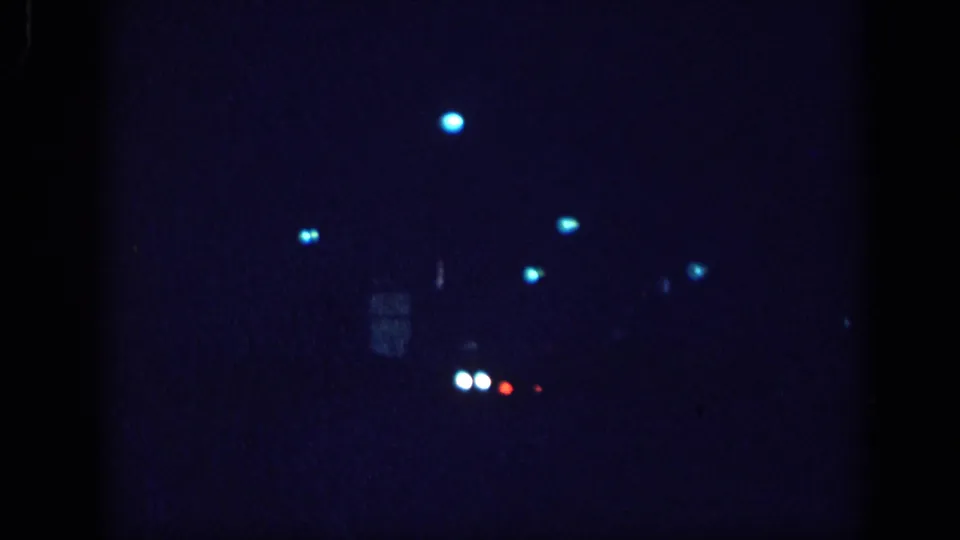8mm home movie night-drive frame, circa 1963 Memphis; paired white bokeh dots suggest an oncoming vehicle's headlights amid scattered blue streetlight bokeh.