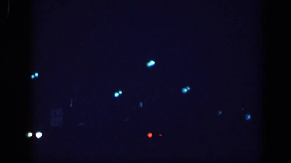 8mm home movie night-drive frame, circa 1963 Memphis; paired white bokeh headlights at bottom-center and scattered cyan streetlight bokeh across a near-black field.