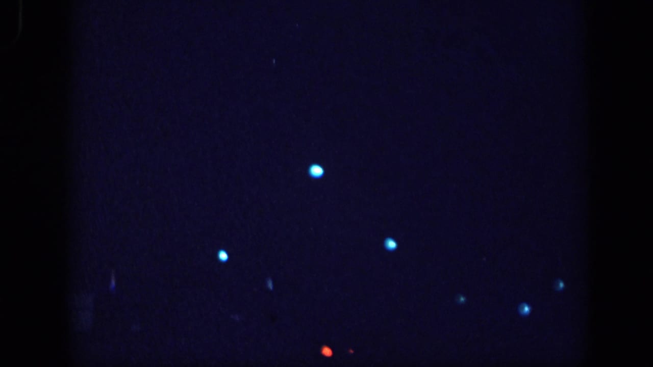 Near-black 8mm film frame showing scattered blue light points and one red artifact, likely film leader or extreme underexposure at the reel opening.