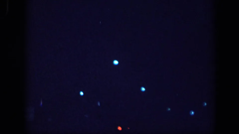 Near-black 8mm film frame showing scattered blue light points and one red artifact, likely film leader or extreme underexposure at the reel opening.
