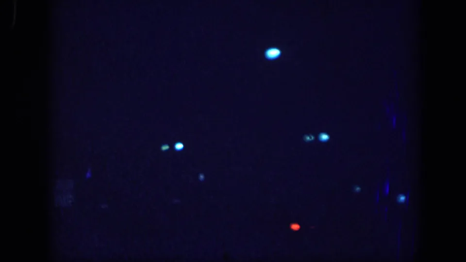 Second dark 8mm frame with denser blue light flares and a single bright circular light at upper center, consistent with film leader before the daytime driving sequence.