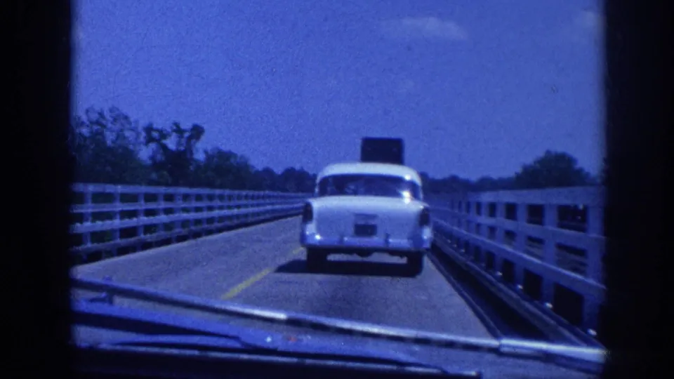 8mm home movie frame showing a white late-model American sedan driving ahead on a bridge with white guardrails and a distant treeline, shot from a following car, Memphis area circa 1963.
