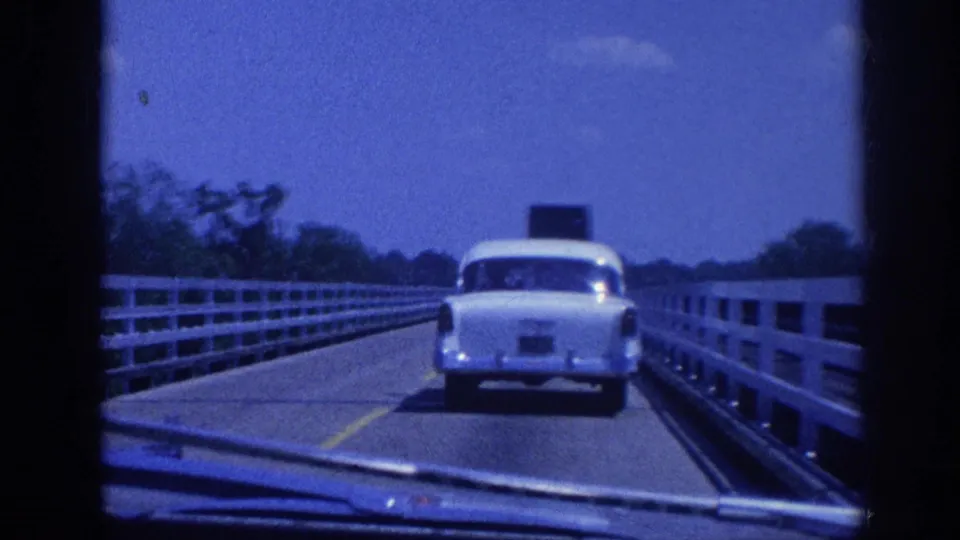8mm home movie frame showing the same white vintage sedan advancing on a bridge with white concrete guardrails and a yellow road marking, treeline on the left bank, circa 1963.
