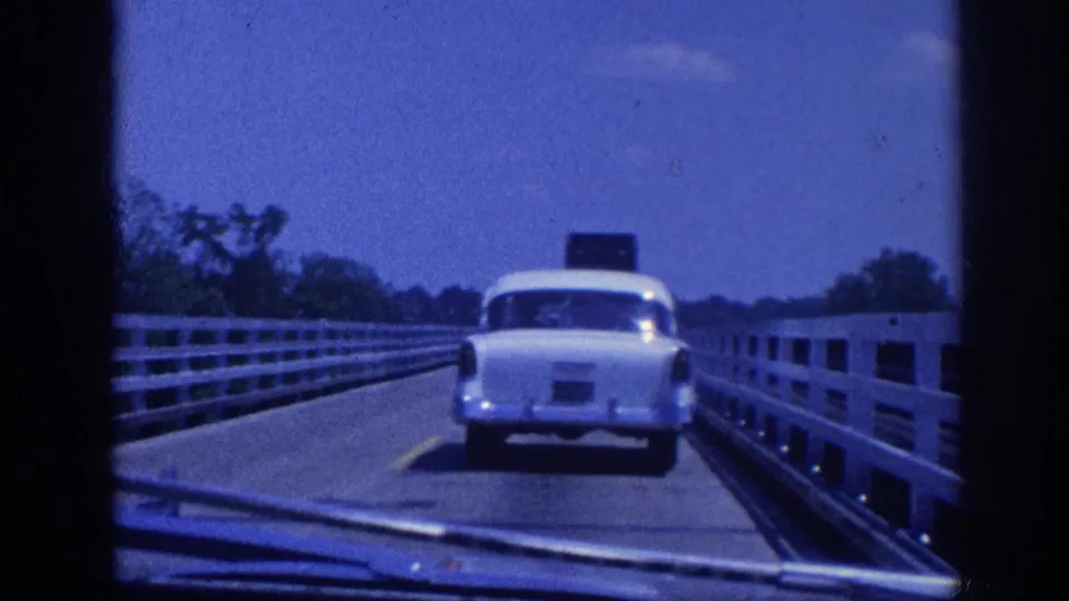 POV from inside a moving car following a late-1950s white sedan across a narrow rural bridge, Memphis Tennessee area, circa 1963, on 8mm film.