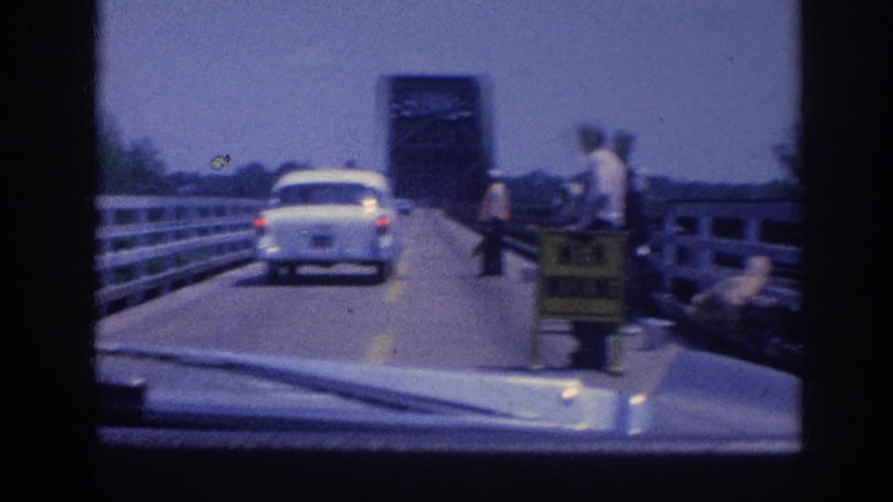 White sedan on a bridge with road workers and a yellow warning sign on the right side; dark steel bridge tower structure visible ahead.