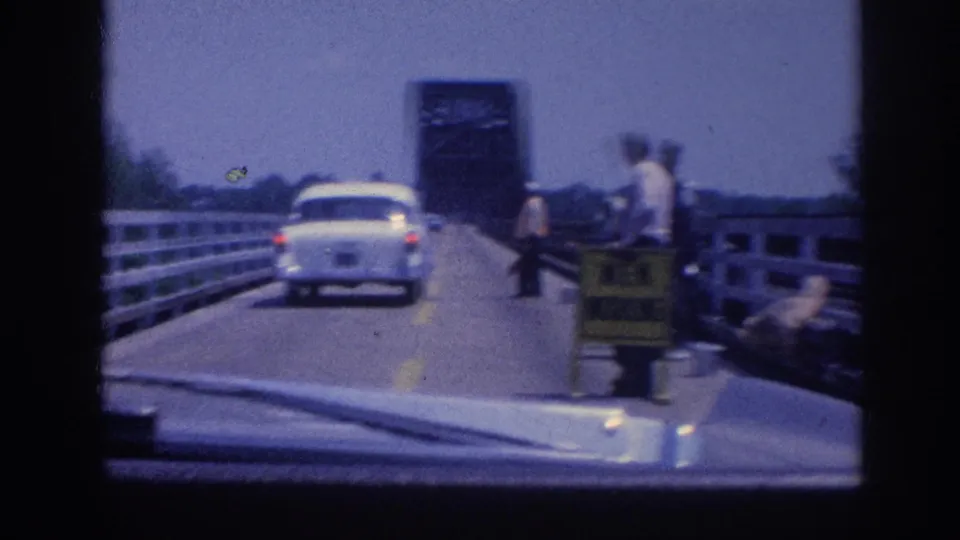 POV following a white sedan on a rural bridge near Memphis; road workers and a yellow warning sign appear on the right, with a steel drawbridge tower ahead, circa 1963.