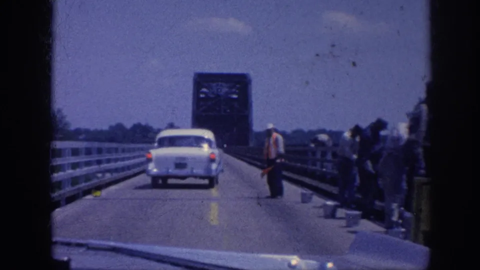 Multiple road workers on both sides of a narrow bridge, one in a bright-colored jacket, with a steel drawbridge tower ahead; Memphis area, circa 1963, 8mm film.