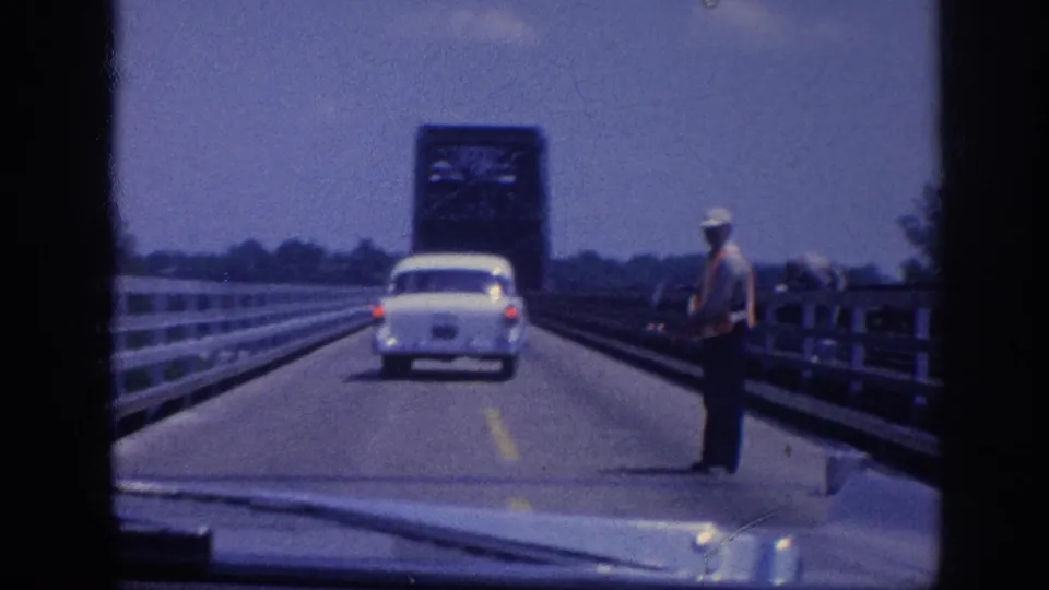 A hatted figure stands at the right bridge edge near a steel drawbridge tower as the white sedan passes through the work zone; Memphis area, circa 1963, 8mm film.