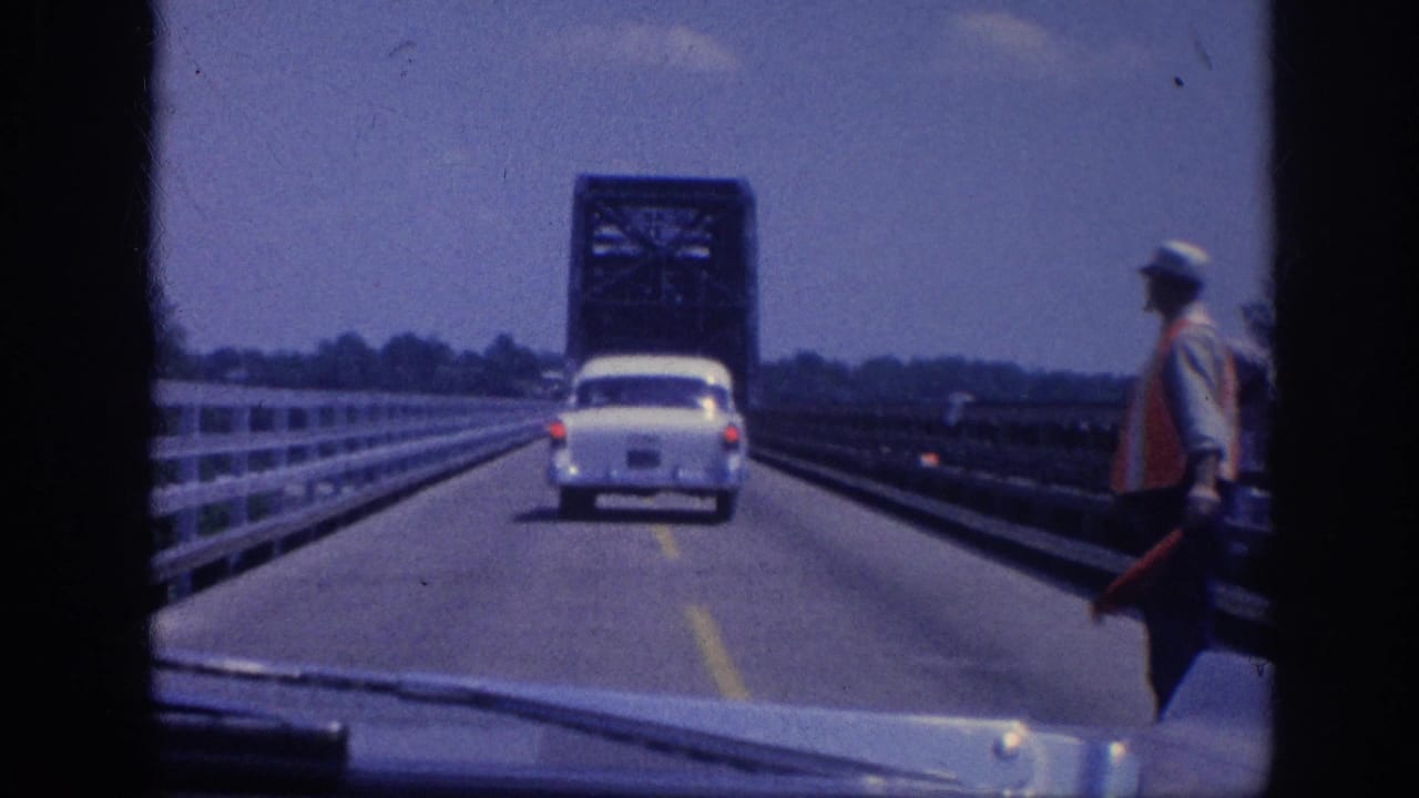 POV from inside a moving vehicle on a steel-truss bridge, circa 1963 Memphis, Tennessee: white American sedan ahead, flagman in orange vest directing traffic, blue sky above.