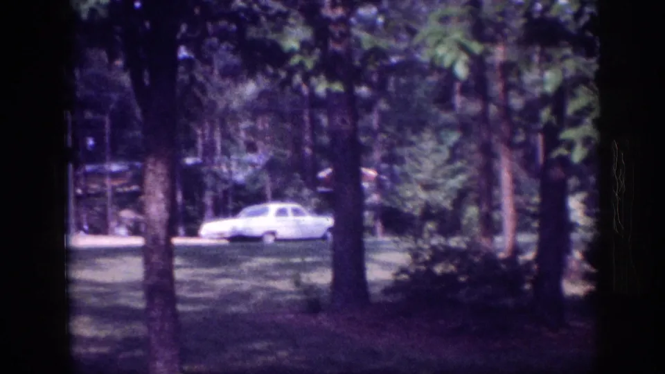 Early-1960s light-colored American sedan visible through tall trees in a wooded clearing, 8mm home movie footage circa 1963.