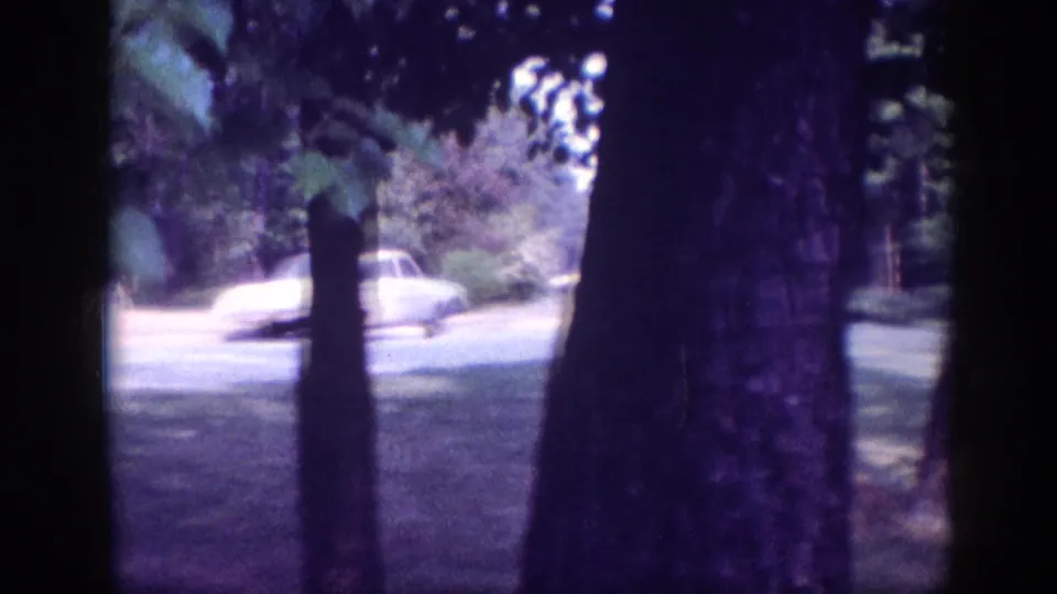White or cream early-1960s American sedan beside a paved driveway in a wooded property, 8mm home movie circa 1963.