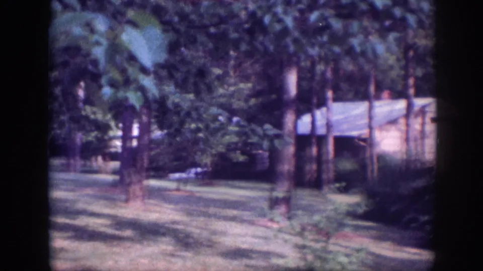 Rustic structure with sloped roof partially visible through trees in a wooded property, 8mm home movie footage circa 1963.
