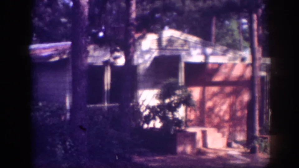 Final pan position reveals brick-toned structure at right and covered shelter amid pines, possibly Memphis Tennessee circa 1963, on aged 8mm film.