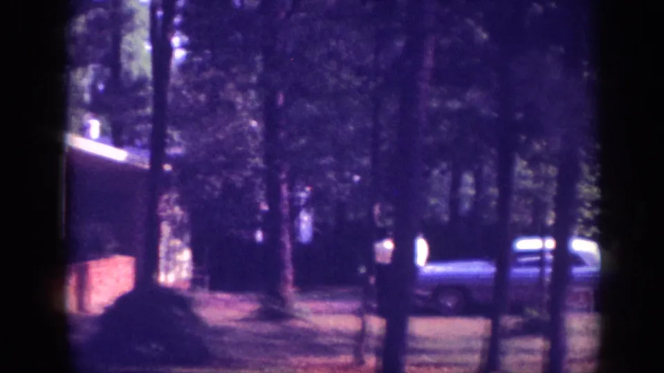 Wooded driveway with a full-size American sedan visible through pine trunks; one or more figures apparent near the car, 8mm footage circa 1963.