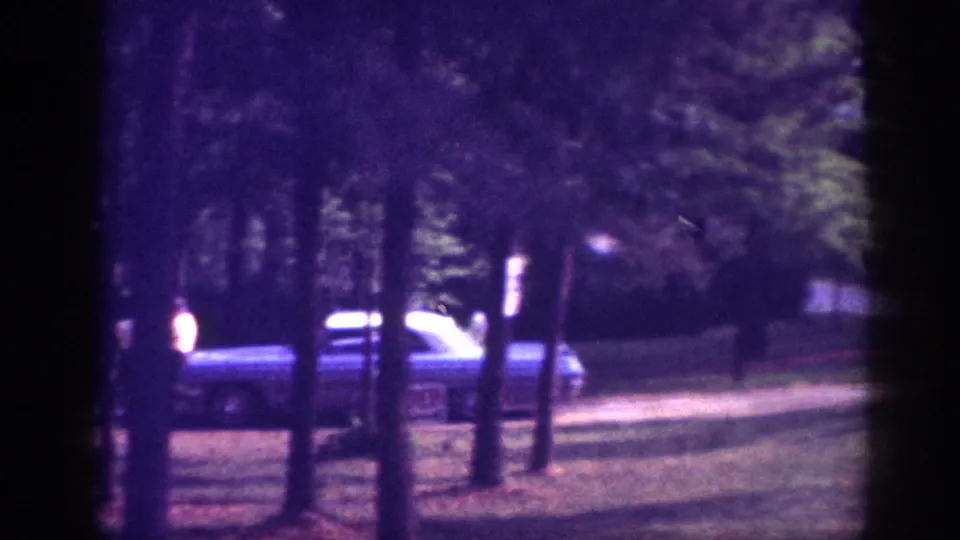 Wooded suburban property with full-size American sedan amid tall pines and a figure near the vehicle, 8mm footage circa 1963.