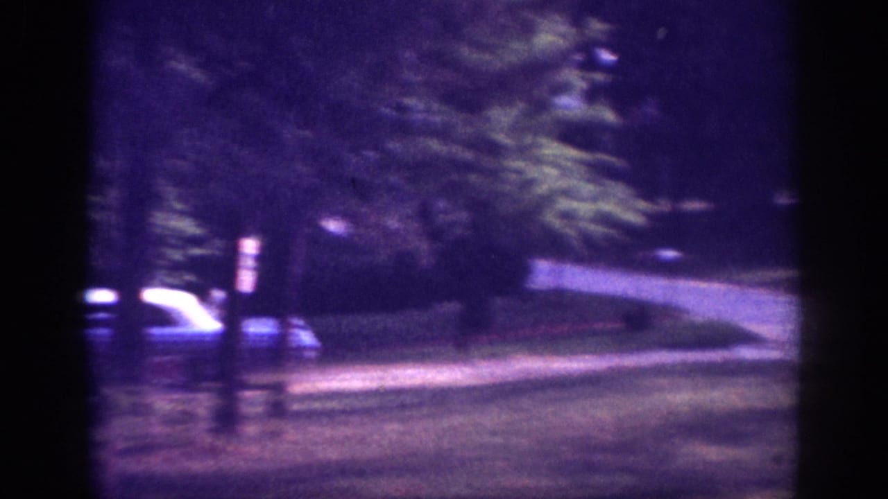 Light-colored vintage automobile near a tree-lined driveway or residential street, shot on aged 8mm film with strong purple color cast and motion blur.