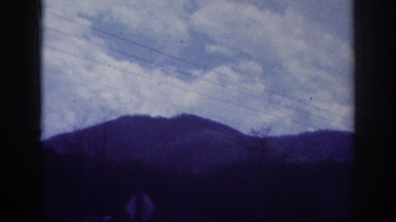 Rolling hills or low mountains under a broad cloudy sky, filmed through a car window on 8mm, with strong blue-purple aging cast; southeastern US road journey, c. 1963.