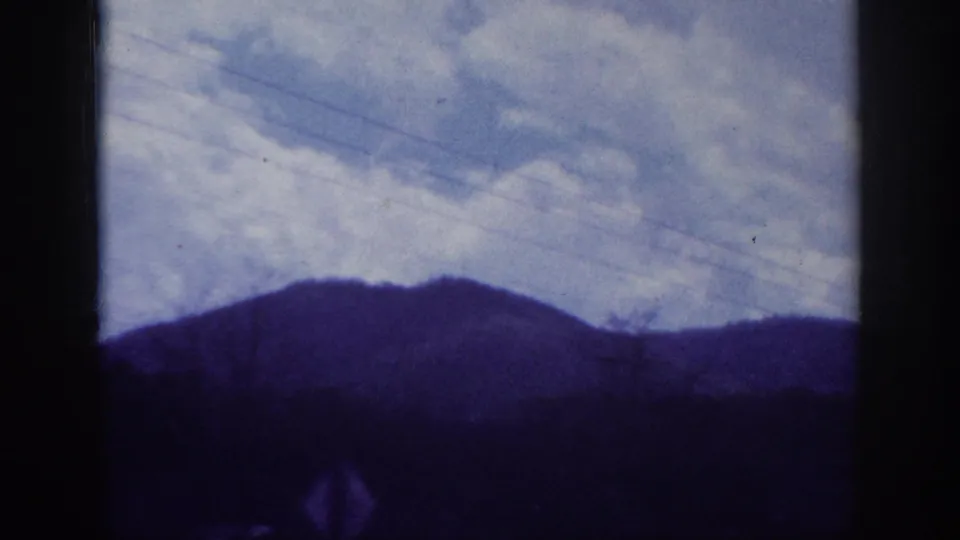 Rolling hills or low mountains under a broad cloudy sky, filmed through a car window on 8mm, with strong blue-purple aging cast; southeastern US road journey, c. 1963.
