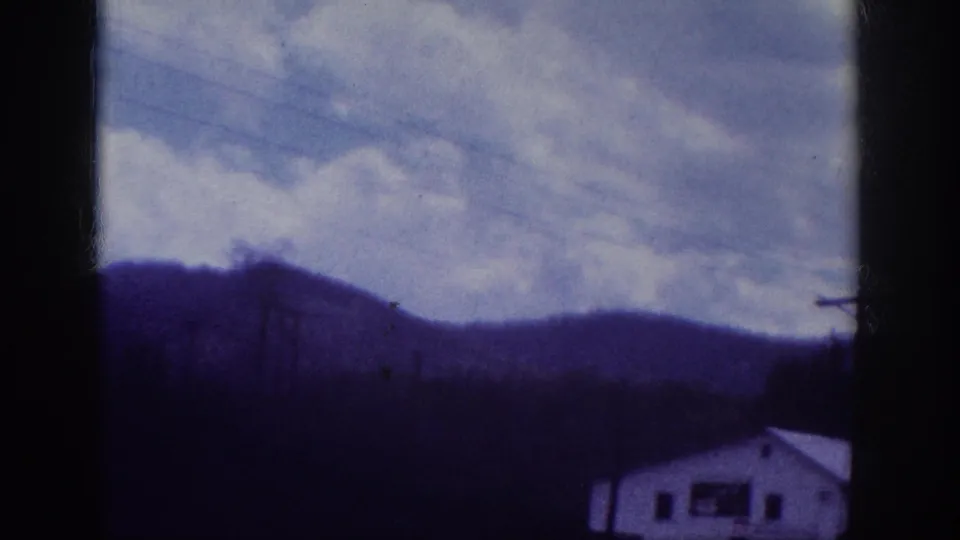 Rural hills, utility lines, utility pole, and a small white building at the frame edge, shot through a car window on 8mm home movie film; southeastern US, c. 1963.