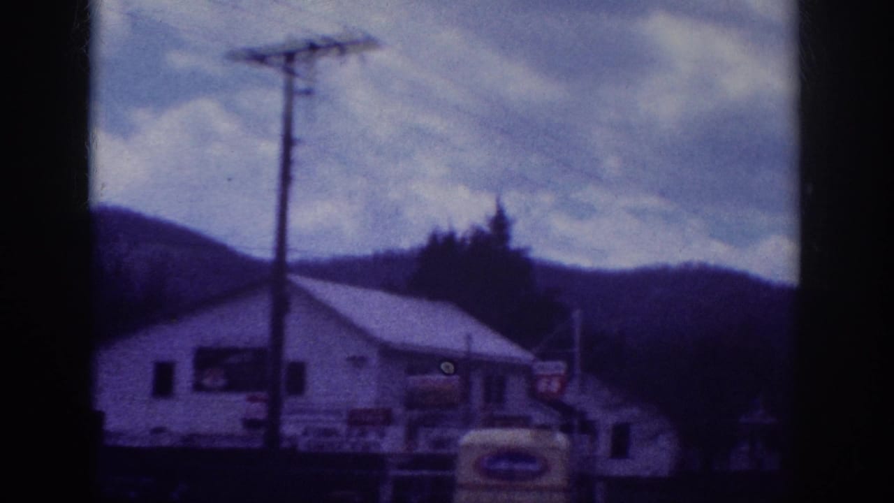 Pan reveals commercial building facade with signage; utility pole centered; partial yellow vehicle at right edge; wooded hills and overcast clouds.