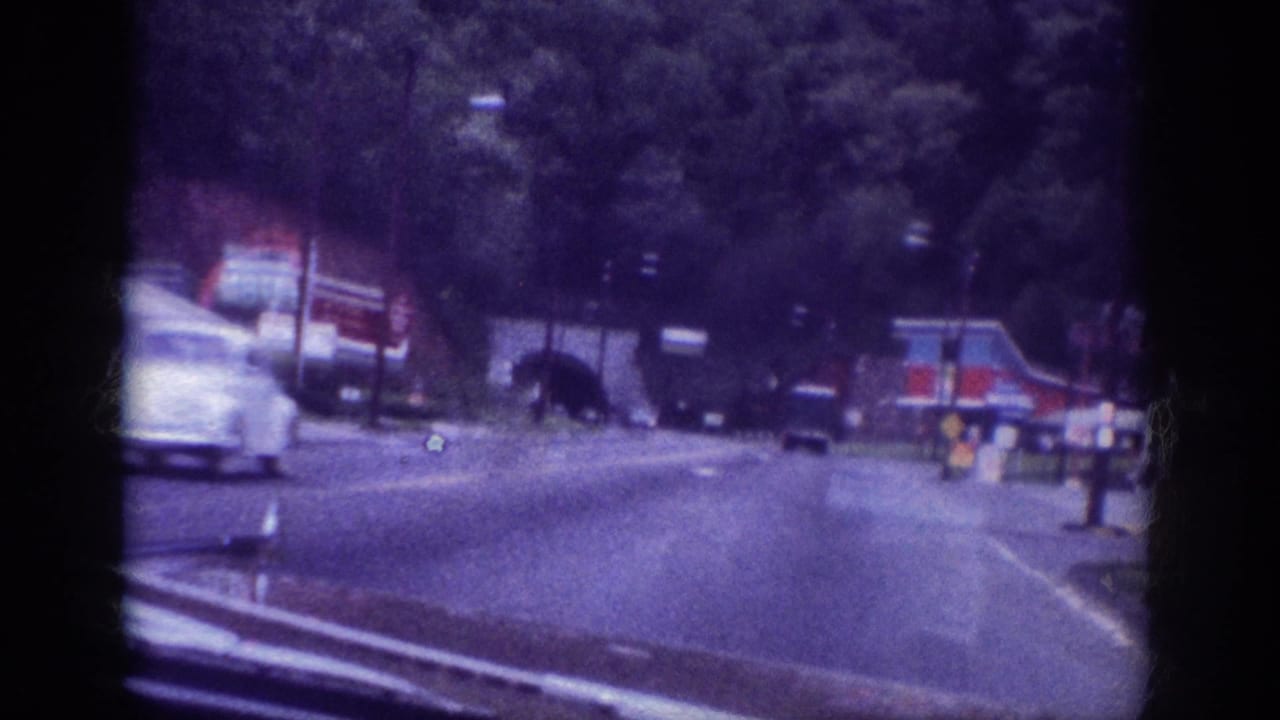 Motion-blurred period automobile passing at speed on a multi-lane road, roadside signs and trees in background, strong blue-purple film cast.