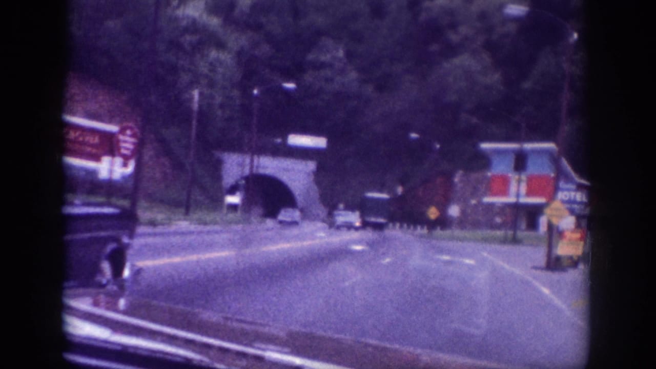 Several 1960s vehicles on a two-lane road approaching a stone-arched tunnel, with roadside commercial signs on the left and trees behind.