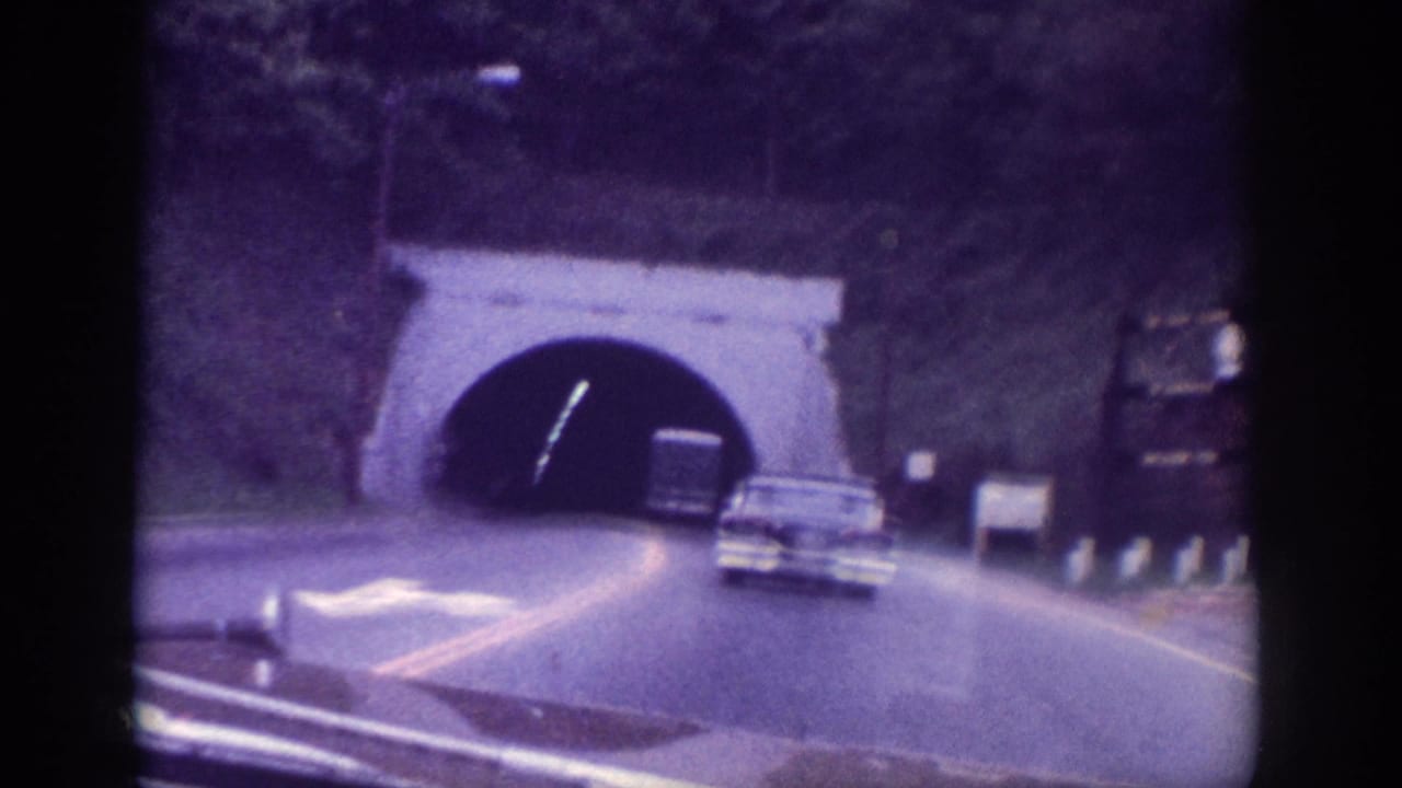 Stone-arched tunnel entrance filling the upper frame, a 1960s sedan on the approach road, white roadside sign at right, trees on hillside above.