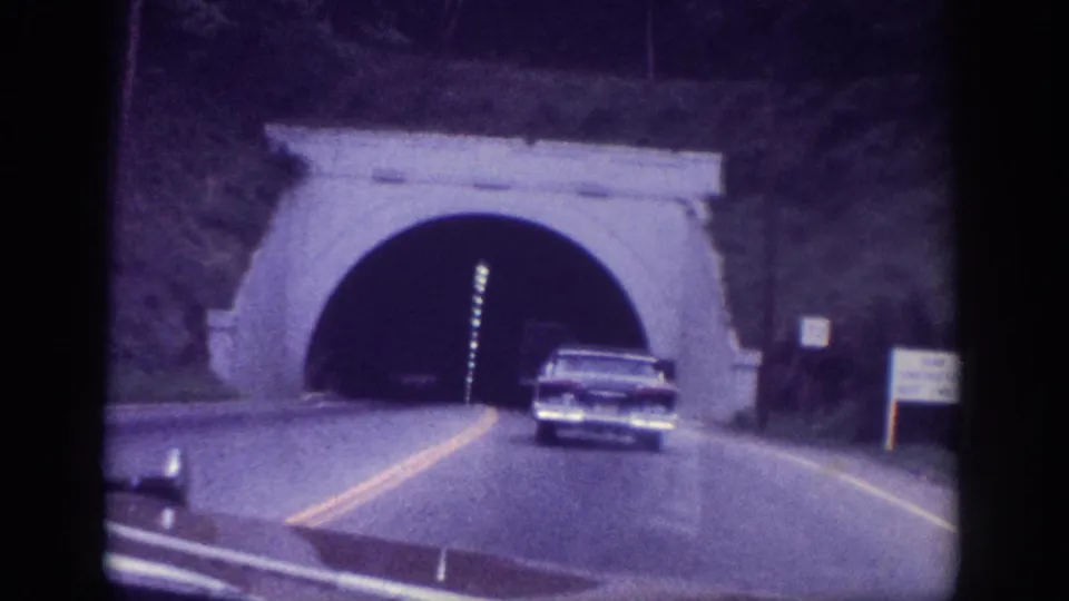 A 1960s large-body American sedan entering a lit stone-arched tunnel, interior ceiling lights visible, shot from a following vehicle on 8mm film, circa 1963.