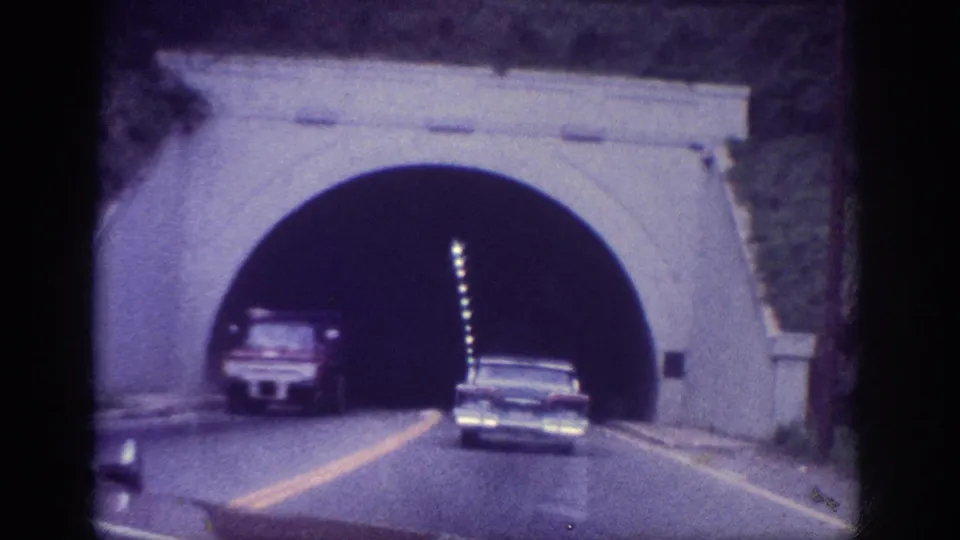 Inside a stone-arched road tunnel, two 1960s American sedans ahead, ceiling lights receding into darkness, shot on 8mm film, circa 1963 Memphis, Tennessee.