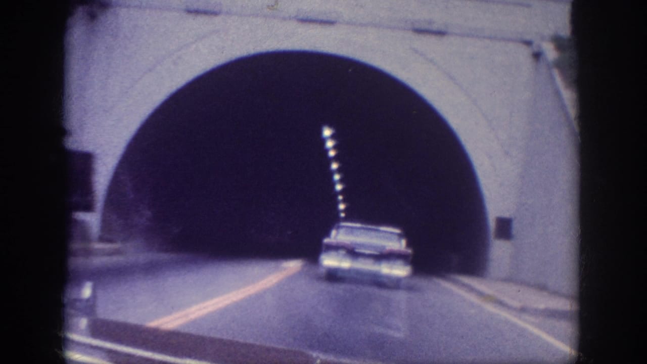 Arched concrete tunnel entrance with a 1960s sedan at the portal and overhead lights receding into darkness.