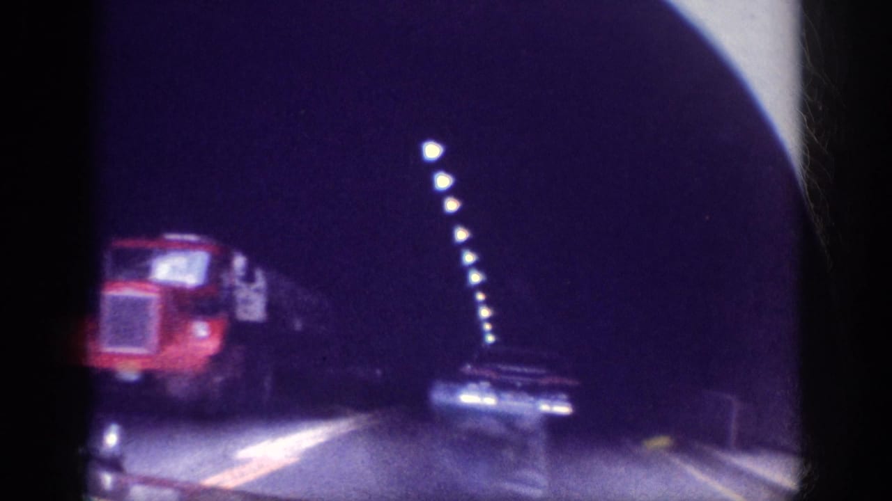 8mm home movie frame circa 1963 showing a red semi-truck ahead inside a road tunnel, overhead lights receding into the darkness with heavy film grain.