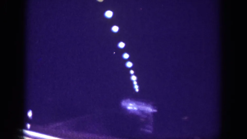 8mm home movie frame circa 1963 deep inside a road tunnel, extreme darkness with overhead lights in a receding diagonal and heavy blue-purple film grain.