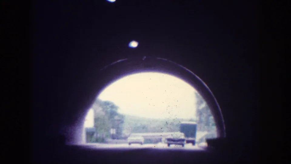 Through the tunnel arch, multiple 1960s-era automobiles are visible on a multi-lane road flanked by trees, filmed on 8mm circa 1963.