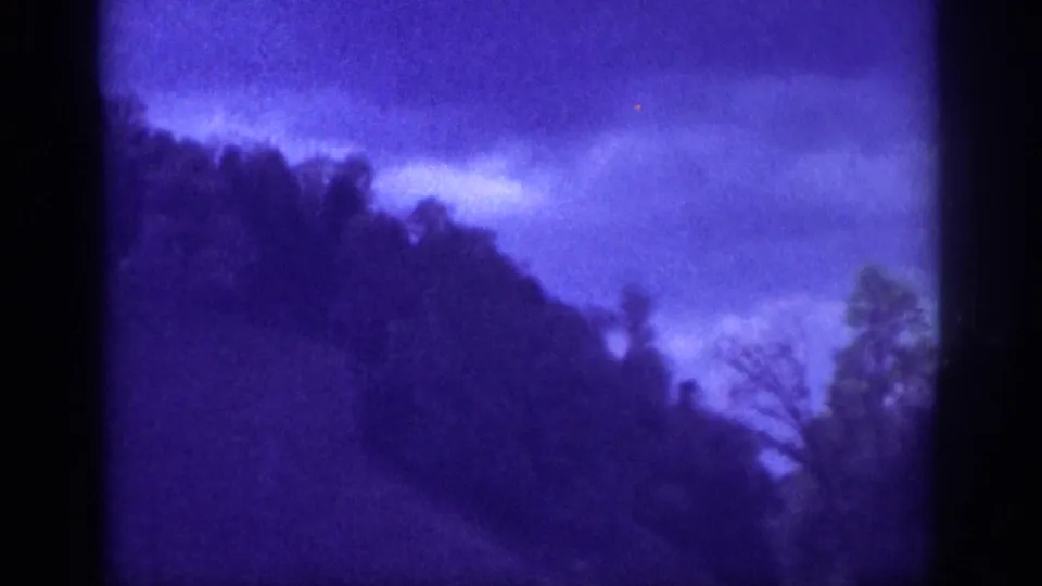 8mm home movie frame circa 1963, passenger-seat view of a winding rural road disappearing into forested hills beneath heavy storm clouds.