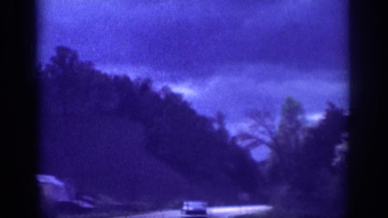 Winding rural road with a dark classic sedan visible in the distance under a stormy blue-purple sky and forested hillside.
