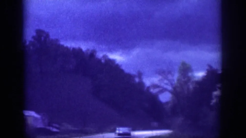 8mm home movie circa 1963, passenger view of a winding rural highway with a classic-era car ahead, stormy overcast sky above forested hills.
