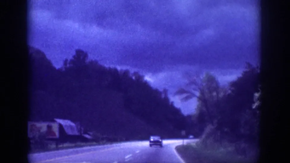 8mm home movie circa 1963, driving view of a two-lane rural highway with a classic car ahead and a partial roadside billboard visible at the left frame edge.