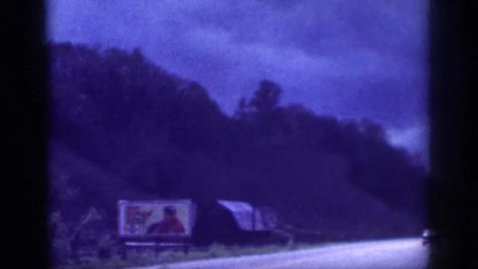 8mm home movie circa 1963, large roadside billboard showing a colorful period-style advertisement alongside a rural American highway; small structure visible to its right.