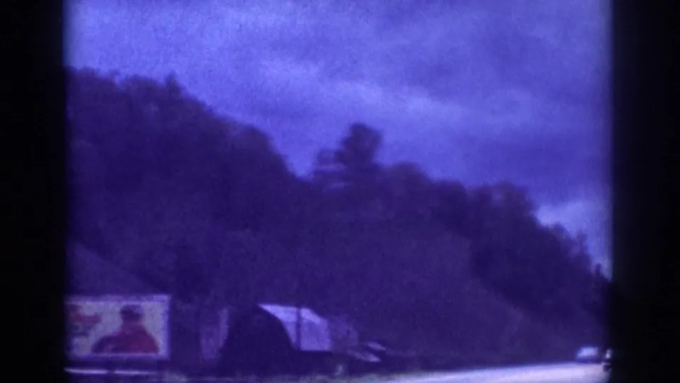 1963 8mm home movie frame — roadside billboard with a depicted figure beside a wooded US highway, heavy purple color cast and film grain throughout.
