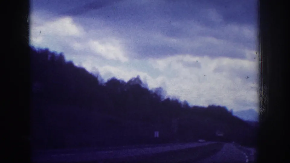 1963 8mm home movie driving shot — wooded hillside flanking a rural US highway under heavy overcast sky, small white roadside sign visible on the right shoulder.
