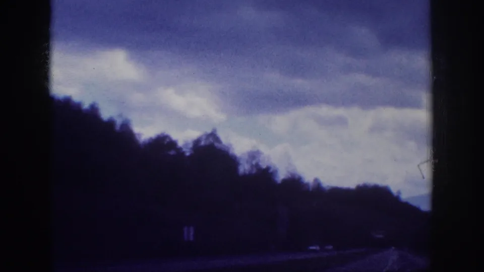 1963 8mm home movie — two-lane highway through wooded terrain, dramatic overcast sky with layered clouds, faint distant vehicles visible on the road.