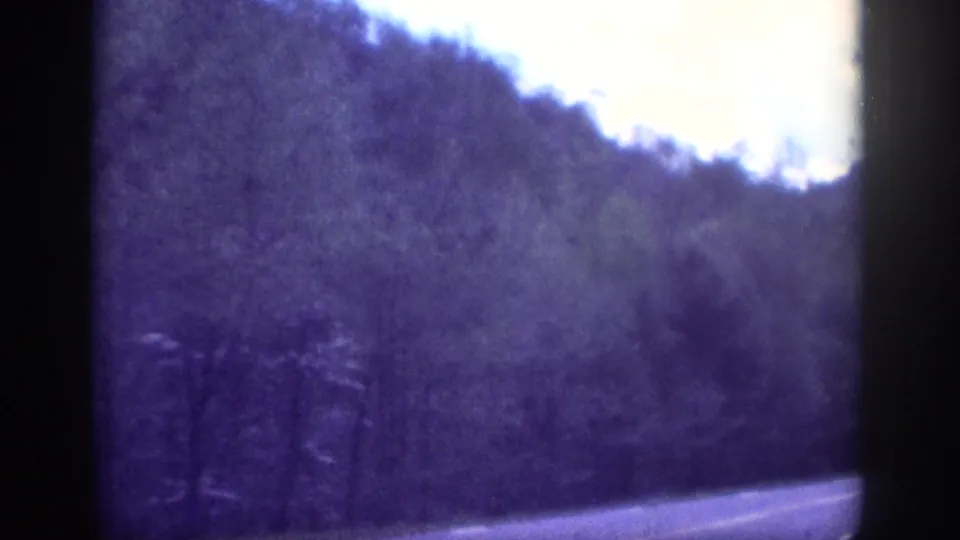 Aged 8mm home-movie frame circa 1963, showing a two-lane road through wooded terrain in the Memphis, Tennessee area under an open sky.