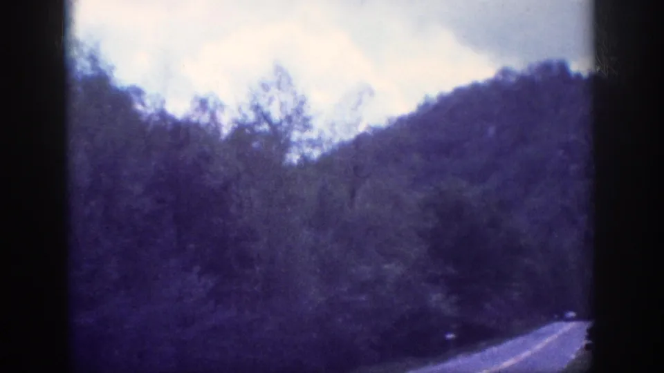 Aged 8mm home-movie frame circa 1963 in the Tennessee area, showing road centre-line markings, a wooded hillside, and partly cloudy sky through a moving vehicle window.
