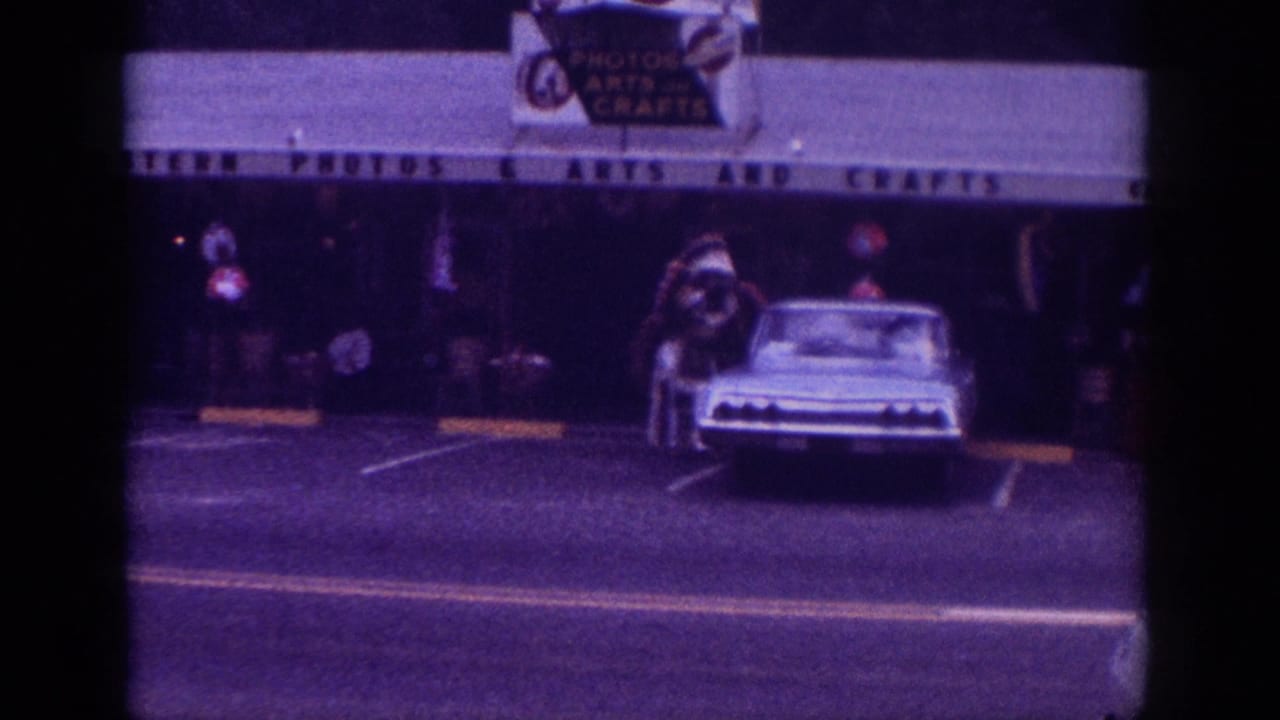 8mm home movie frame, likely Memphis Tennessee c.1963, showing a light-colored American sedan and costumed street figure outside an arts and crafts store, purple color cast.