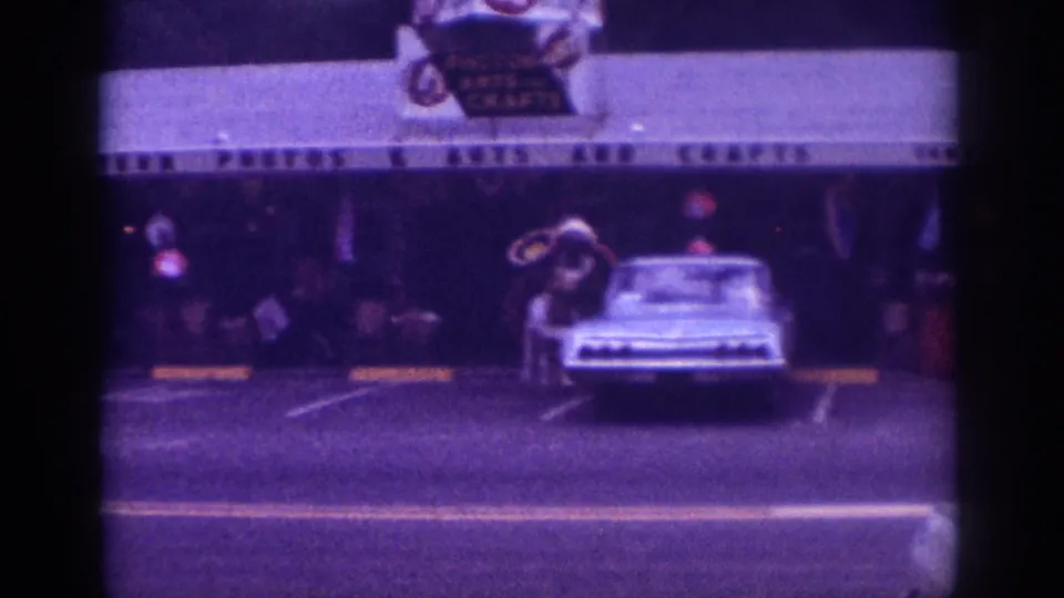 8mm home movie, Memphis Tennessee c.1963, light-colored American sedan and costumed figure with circular props outside 'Photos & Arts and Crafts,' purple-tinted aged film stock.